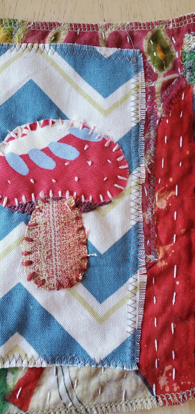 May include: A textile artwork with a red mushroom appliqu&eacute;, white stitching, and a blue and white chevron pattern. The mushroom has a woven stem. Other fabric patterns form the background.