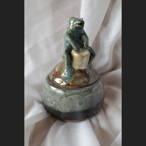 May include: A ceramic jar with a lid shaped like a frog playing a drum. The jar is glazed in shades of brown, green, and black.