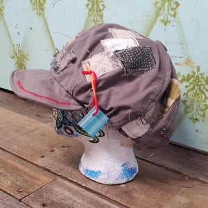 May include: Grey patchwork cap with a red stitched brim, displayed on a white mannequin head. The cap features various fabric patches in neutral tones. A small tag hangs from the cap.