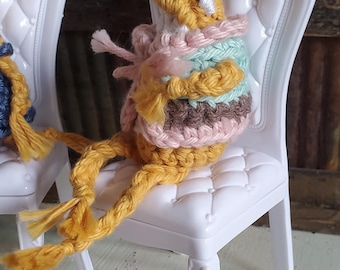 Può includere: Una creatura fatta a mano, all'uncinetto con filato giallo, rosa e blu, è seduta su una piccola sedia bianca. La creatura ha lunghe gambe di filato giallo e indossa un maglione multicolore. La sedia ha uno schienale decorativo ed è posizionata su una superficie verniciata.