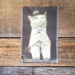 May include: A monochrome image of a cat in a pinstripe suit, bow tie, and top hat. The cat is standing, gazing upwards. The image is on a rectangular fabric with a frayed edge, set against a wooden background.