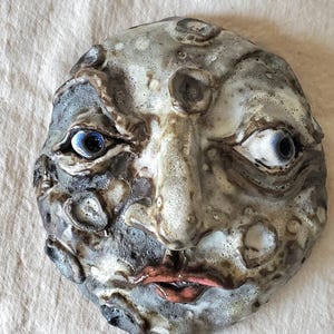 May include: A ceramic face sculpture with a textured, aged appearance. The face has blue eyes, a prominent nose, and red lips. The sculpture is primarily gray and white with dark brown accents, and is set against a neutral background.