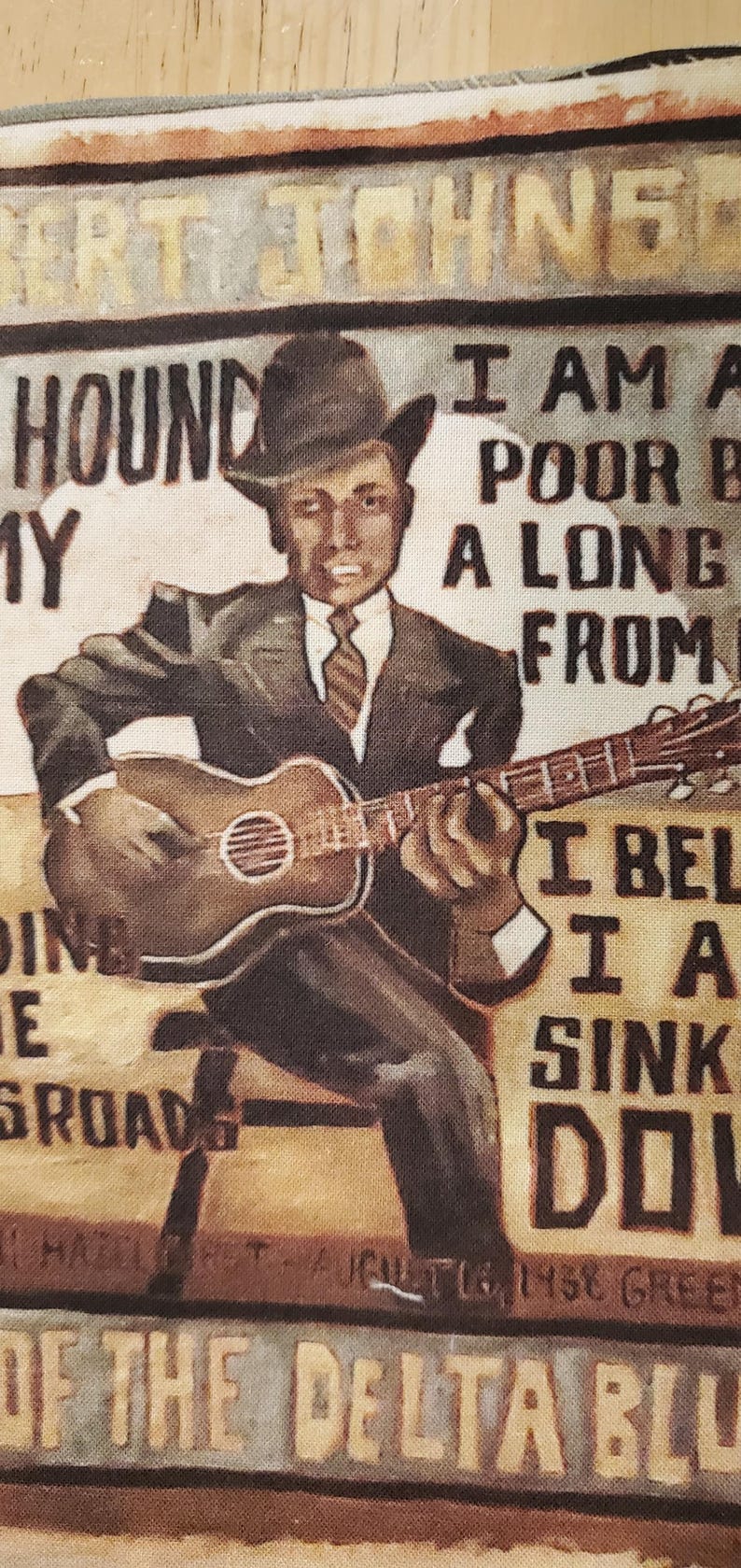 Pu&ograve; includere: Pannello in tessuto con un'illustrazione vintage di un uomo che suona la chitarra. L'immagine include il testo "Robert Johnson", "Hound My" e "Of The Delta Blues". L'uomo indossa un abito e un cappello.