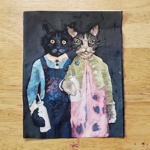 Op de afbeelding: Een zwarte kat en een grijs-witte kat die kleding dragen en melkflessen met rietjes vasthouden. De zwarte kat draagt een blauw shirt en een overall. De grijs-witte kat draagt een groene trui en een roze jurk met blauwe bloemen. De achtergrond is donkergrijs.