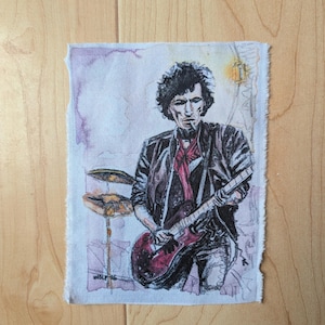 May include: A rectangular fabric patch featuring a detailed ink and watercolor illustration of a musician with dark curly hair, wearing a black leather jacket and a red scarf, playing a red electric guitar, with cymbals visible in the background.