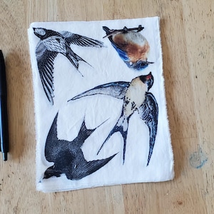 May include: A white fabric square with four bird illustrations. The birds are depicted in various poses, with detailed black and white wings and bodies. One bird has blue and brown coloring. The fabric has a raw edge.