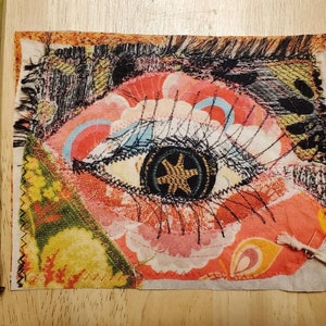 May include: A colorful fabric patch with an embroidered eye design. The eye is surrounded by pink, yellow, and blue floral patterns. The fabric is textured and has a frayed edge.