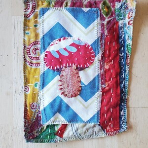 May include: A handmade fabric art piece featuring a red mushroom applique on a blue and white chevron background. The artwork is framed by colorful patterned fabrics and hand-stitched details, showcasing a rustic, bohemian aesthetic.