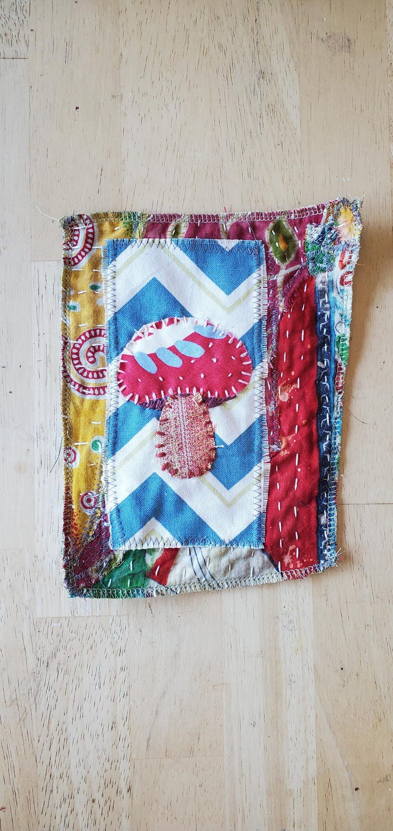 May include: A patchwork textile art piece with a red mushroom design on a blue and white zigzag pattern. The artwork is surrounded by various patterned fabrics and hand-stitched details, displaying a rustic, bohemian style.