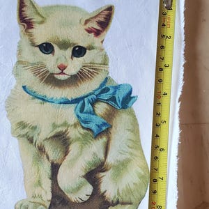 May include: A vintage-style fabric square featuring a seated kitten with a blue bow. The kitten has light yellow fur, blue eyes, and a pink nose. The fabric measures approximately 6 inches in length.