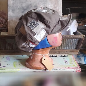 May include: A brown newsboy cap with fabric patches sewn on. The patches have different textures and patterns, giving a unique, handcrafted appearance. The cap is displayed on a mannequin head with a blue neck and a blank tag.