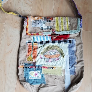 May include: A beige tote bag with a patchwork design. The bag features a long strap with colorful fabric patterns. Patches of various colours and textures are sewn onto the front, including an eye design. The text "THE WOODEN WOLF" is visible.