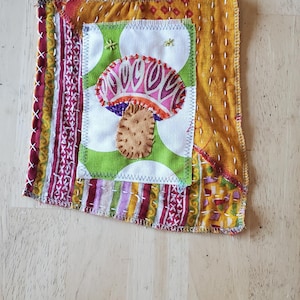 May include: A handmade fabric square with a mushroom applique. The mushroom has a pink and purple cap and a brown stem. The square is made of various fabrics in shades of yellow, red, and green, with decorative stitching.