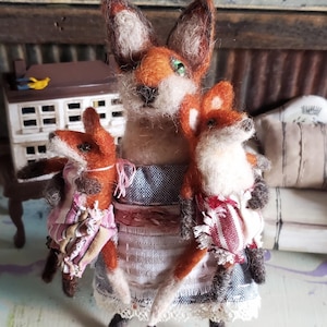 May include: A felt fox family figurine. The mother fox wears a blue and white dress with a lace trim, holding two smaller foxes. The foxes have orange fur, white faces, and green eyes. The background includes a small house and decorative elements.