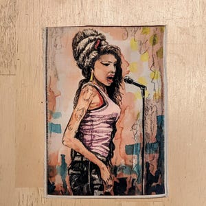 May include: A rectangular fabric patch featuring a watercolor-style illustration of a singer with a high beehive hairstyle, wearing a light pink tank top and dark pants, standing at a microphone stand against a peach and blue abstract background.