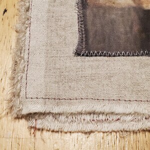 May include: Close-up of a fabric piece with a printed image, featuring a frayed edge and a stitched border. The fabric has a natural, beige tone, with a detailed image on the right side. The edges are finished with a red stitch.