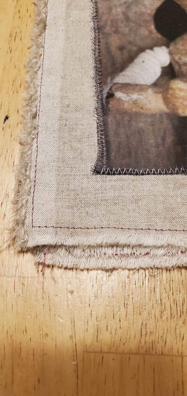 May include: Close-up of a fabric piece with a printed image, featuring a frayed edge and a stitched border. The fabric has a natural, beige tone, with a detailed image on the right side. The edges are finished with a red stitch.