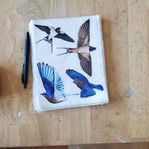 May include: A white square fabric with bird illustrations. The birds include swallows and bluebirds, in shades of blue, black, white, and brown. A black pen lies to the left of the fabric. The fabric is on a wooden surface.
