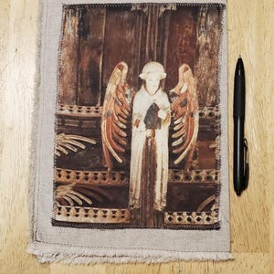 May include: A linen fabric panel with a printed image of a white angel with outstretched wings. The angel is holding an object, set against a brown background. The panel is framed with a frayed edge. A black pen is next to the panel.