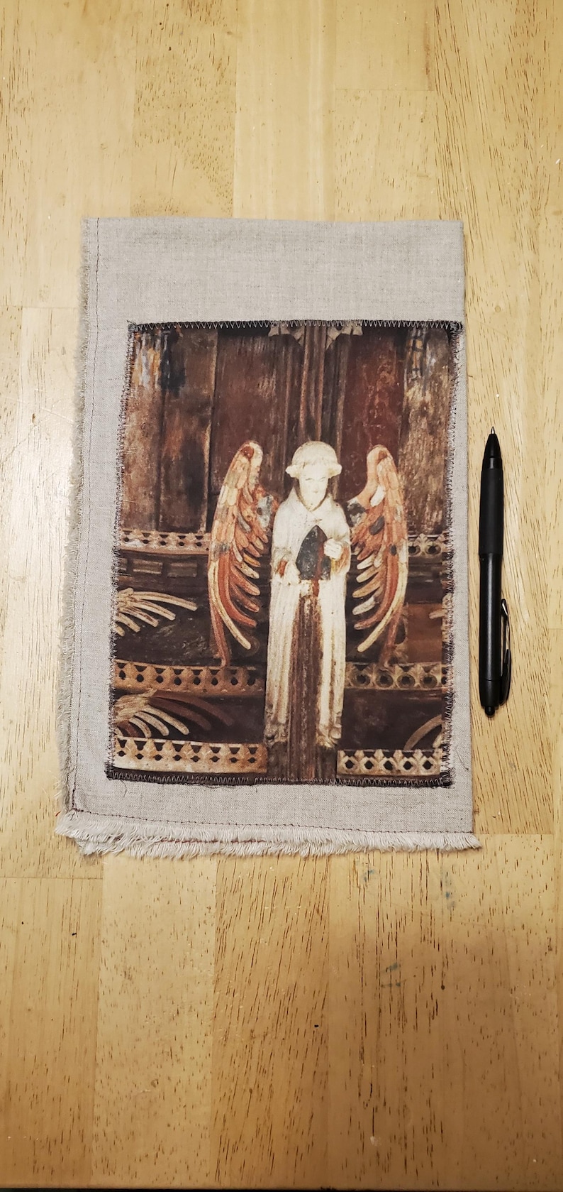 May include: A linen fabric panel with a printed image of a white angel with outstretched wings. The angel is holding an object, set against a brown background. The panel is framed with a frayed edge. A black pen is next to the panel.