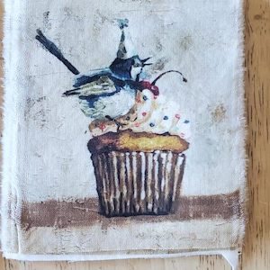 May include: Square fabric with a whimsical illustration of a blue bird perched above a cupcake with white frosting, red cherry, and colorful sprinkles. The cupcake has a brown striped wrapper. The fabric has a distressed, vintage look.