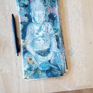 May include: A stack of fabric with a detailed illustration of a seated Buddha in shades of blue, green, and white, surrounded by floral elements. A black pen rests beside the fabric on a wooden surface.
