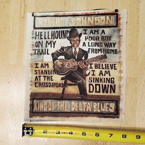 Pu&ograve; includere: Un'opera d'arte quadrata in tessuto con un dipinto di Robert Johnson che suona la chitarra. Il testo include "Hellhound on my Trail" e "King of the Delta Blues". Il pezzo misura circa 15 cm di larghezza.