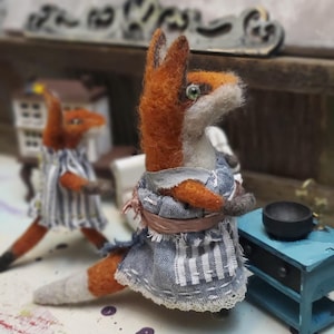 May include: Needle felted fox figurine wearing a blue and white striped apron and dress. The fox has orange fur, a white muzzle, and green eyes. A second fox figurine is in the background. A small blue table and black bowl are also visible.