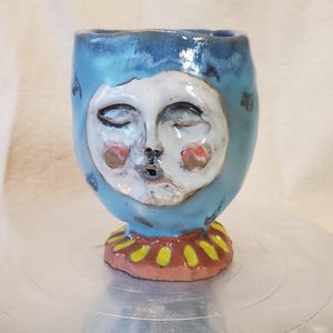 May include: A handmade ceramic cup with a whimsical face design. The cup is primarily blue with a white face featuring closed eyes, rosy cheeks, and a small mouth. The base is decorated with yellow and red accents.