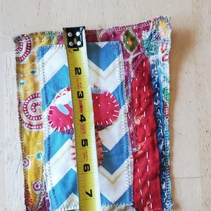 May include: A colourful, handmade fabric square with a patchwork design. The square features various patterns, including a blue and white chevron, red strawberry shapes, and yellow floral fabric. The square is approximately 20 cm long.
