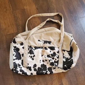 May include: A beige and black cow print duffel bag with two handles. The bag has a rectangular shape with a large main compartment and a smaller front pocket. The bag is made of canvas material.