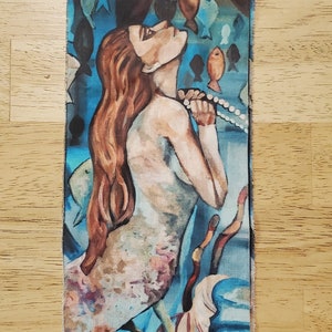 May include: A painting of a mermaid with long red hair, wearing a pearl necklace, surrounded by blue and orange fish. The mermaid is looking up and has a serene expression. The background is a vibrant blue with hints of green and white.