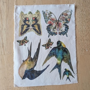 May include: A rectangular piece of off-white fabric featuring printed illustrations of two butterflies and three birds. The top butterfly has yellow and black wings, while the other has floral patterns. Two swallows are depicted in flight alongside a smaller bird.