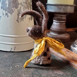 May include: A brown felt rabbit with a yellow ribbon around its neck. The rabbit is standing on a wooden surface.