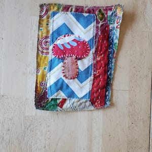 May include: A handcrafted fabric patch with a red mushroom design on a blue and white zigzag pattern. The patch is made from various patterned fabrics and stitched with visible thread.