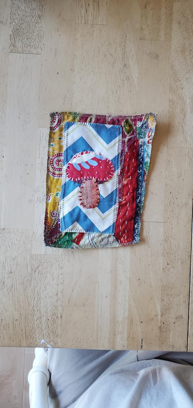 May include: A handcrafted fabric patch with a red mushroom design on a blue and white zigzag pattern. The patch is made from various patterned fabrics and stitched with visible thread.