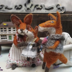 May include: A felted fox family, featuring three orange foxes. One fox wears a dress with a floral and striped pattern, another wears a dress with musical notes. The third fox carries a grey bundle. The foxes are on a white surface.