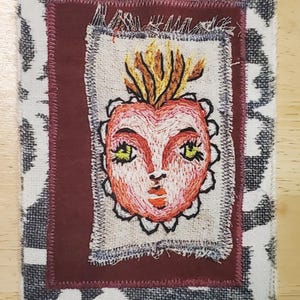 May include: Embroidered textile art piece featuring a heart-shaped face with green eyes and orange lips, surrounded by a flame-like design. The artwork is set within a patchwork of fabrics, including a patterned border.