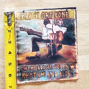 May include: A vintage-style patch featuring a black man playing a guitar, with the text "BLACK CAT BONE SWAMP BOOGIE QUEEN RHYTHM AND BLUES" and a gold star.