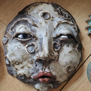 May include: A ceramic face sculpture with a textured, aged appearance. The face has blue eyes, a slightly crooked mouth, and a detailed, expressive design. The sculpture is in shades of gray, brown, and pink.