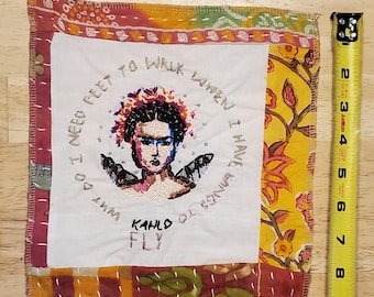 Può includere: Arte tessile ricamata raffigurante Frida Kahlo con la scritta "Perché ho bisogno di piedi per camminare quando ho le ali per volare?" e "Kahlo Fly". L'opera è incorniciata con tessuto fantasia colorato.
