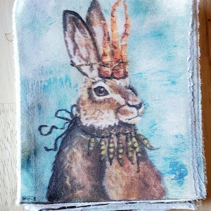 May include: A brown rabbit with a crown of antlers and a green and yellow beaded necklace on a blue background. The rabbit is looking to the right. The image is printed on a white fabric.