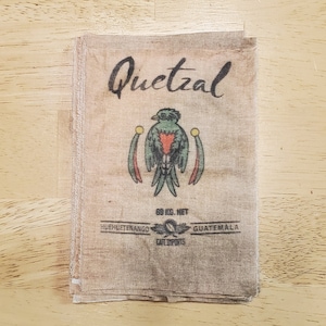 May include: A stack of fabric squares with a printed design featuring the word "Quetzal" in script above a colorful bird illustration. The text "60 KG. NET" and "HUEHUETENANGO GUATEMALA CAFE IMPORTS" are also visible.