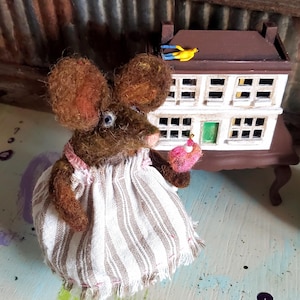 May include: A handmade mouse figure with brown fur, wearing a striped dress and holding a pink cupcake. A miniature house with a brown roof and white walls is in the background. The scene is set on a painted surface.