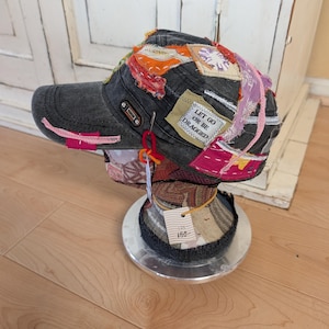 May include: A charcoal grey baseball cap featuring various colourful fabric patches, frayed edges, and visible stitching. A rectangular patch on the side displays the text "LET GO OR BE DRAGGED" in black lettering. The cap is displayed on a mannequin head.