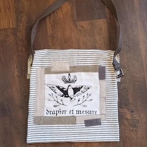May include: A striped tote bag with a brown leather strap. The bag has a central patch with a bird and crown design, and the text "drapier et mesure".