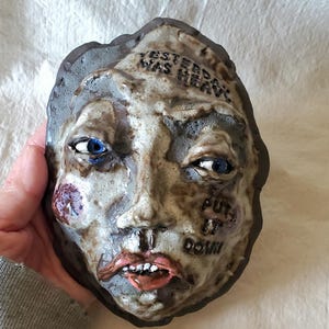 May include: A ceramic face sculpture with blue eyes, red cheeks, and open mouth. The words "YESTERDAY WAS HEAVY" and "PUT IT DOWN" are inscribed on the sculpture. The sculpture is held by a person.