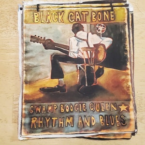 May include: A vintage-style artwork featuring a musician playing a guitar, with the text "BLACK CAT BONE" and "SWAMP BOOGIE QUEEN RHYTHM AND BLUES". The artwork has a distressed, aged appearance, with a warm color palette.