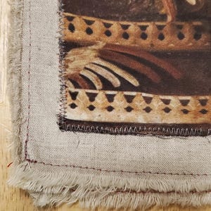 May include: Close-up of a fabric square with a printed design featuring brown and tan tones. The design includes geometric shapes and stylized figures. The fabric has a frayed edge and is sewn with red thread.