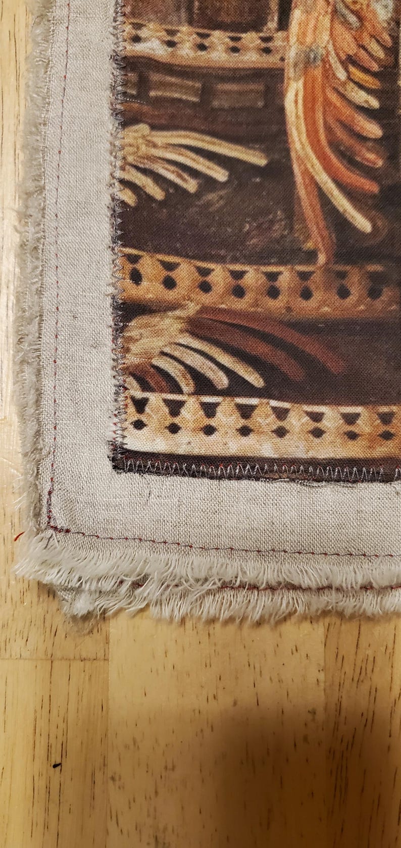May include: Close-up of a fabric square with a printed design featuring brown and tan tones. The design includes geometric shapes and stylized figures. The fabric has a frayed edge and is sewn with red thread.